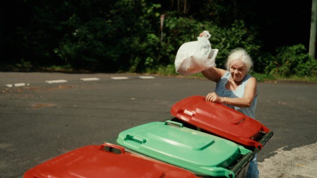 Photograph of an elderly woman putting some rubbish in a wheelie bin.